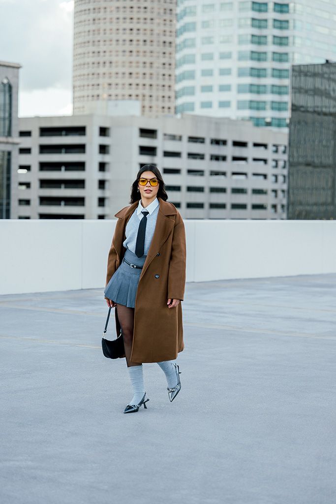 Fashion-forward woman in brown coat, gray pleated skirt, tie, and orange glasses walks on urban rooftop.