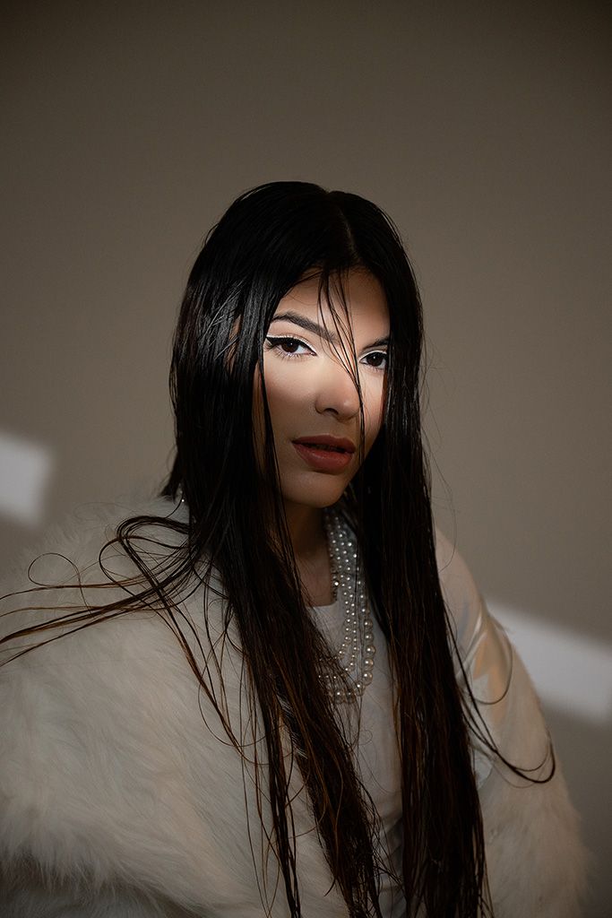 A woman with long black hair wearing a pearl necklace and white fur coat poses elegantly against a neutral background.