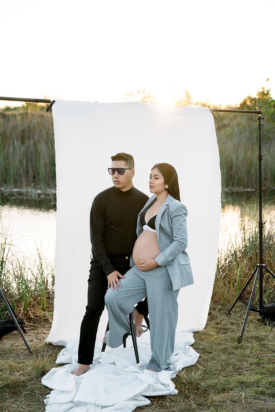 Maternity photo shoot by a lake at sunset with white backdrop and professional lighting setup.