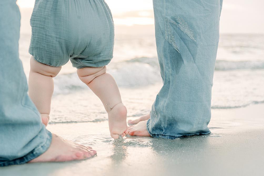 Baby taking first steps on sandy beach with parent, waves gently washing over their bare feet in shallow water.