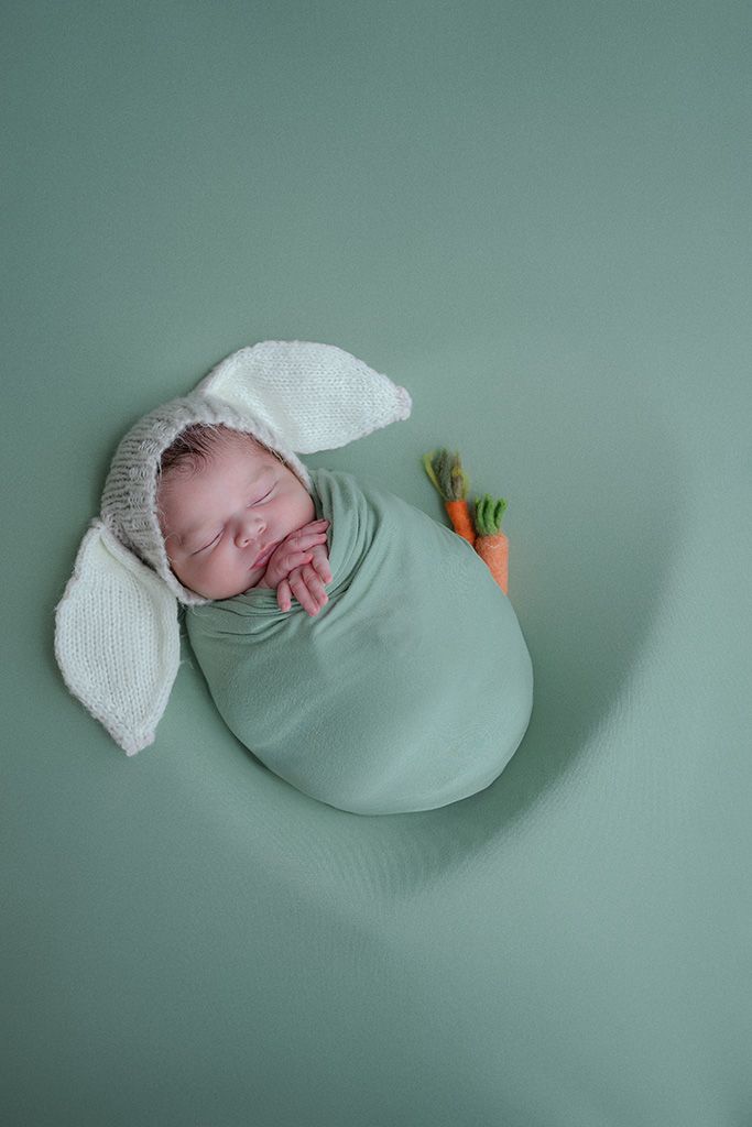 Sleeping newborn baby swaddled in green wrap wearing bunny ear hat with tiny carrot prop on sage green background.