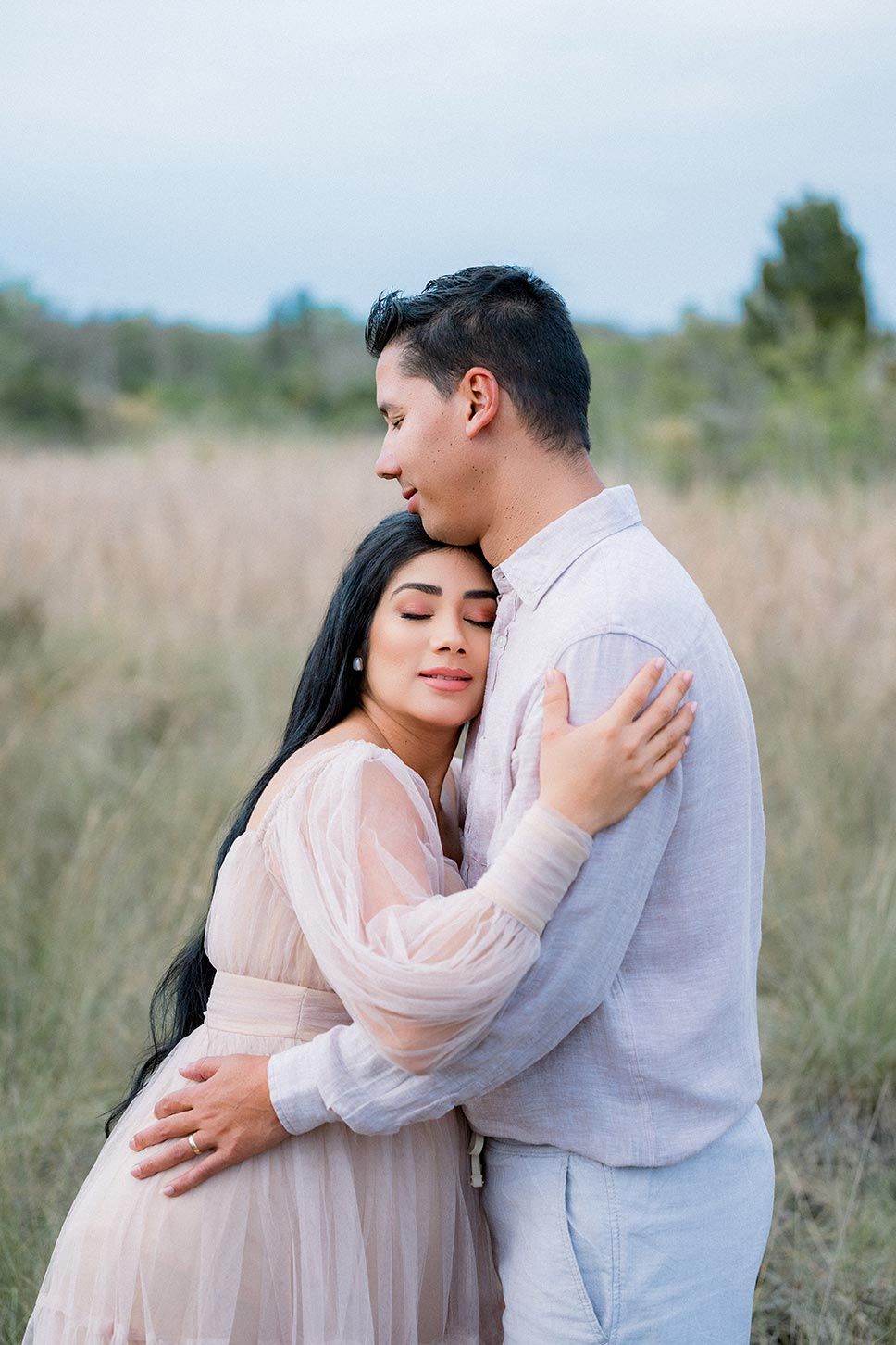 A couple embraces tenderly in a golden field, the man kissing her head as she closes her eyes in a blush tulle dress.