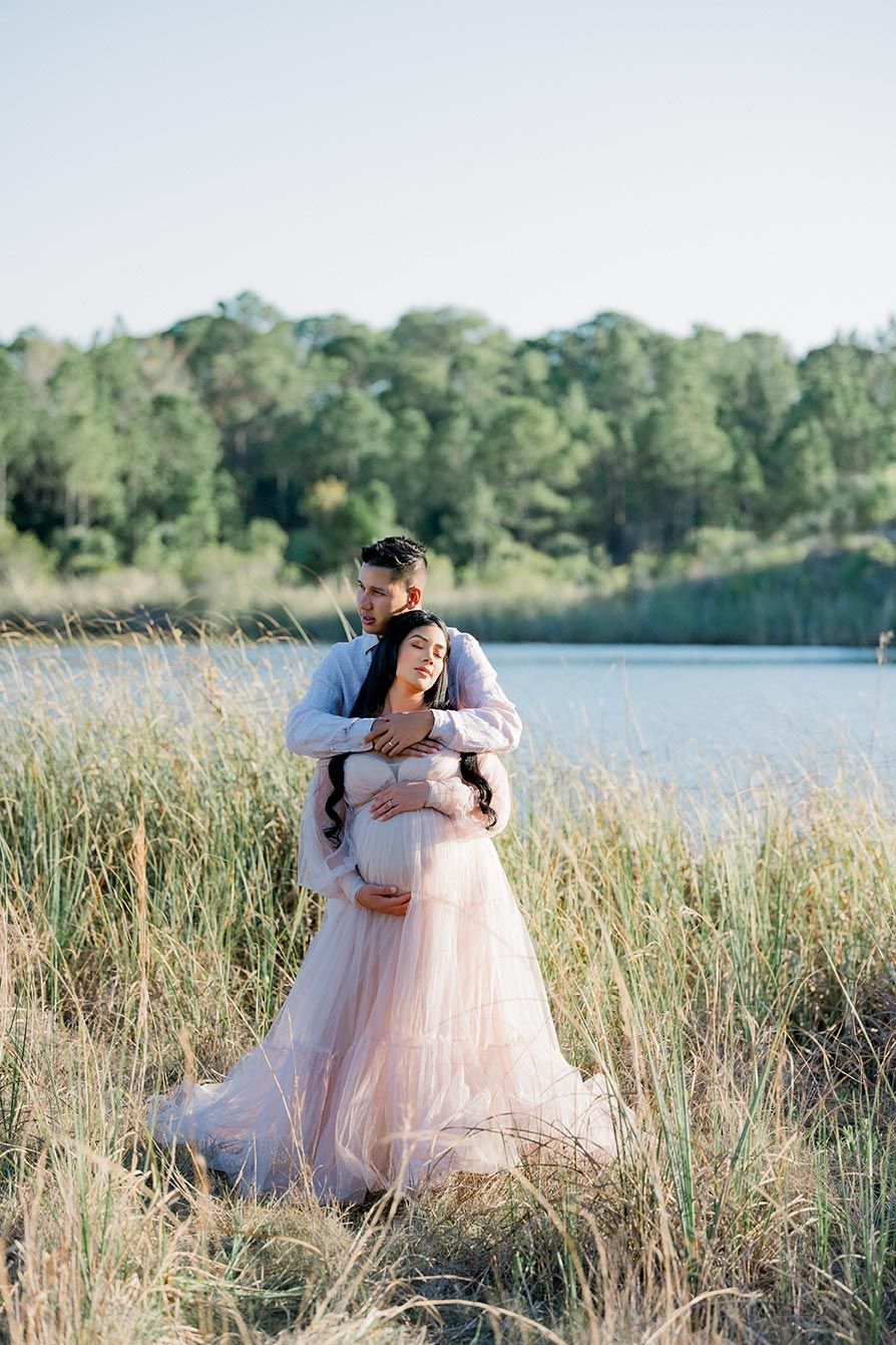 A couple shares an intimate moment in tall grass by a lake at sunset in soft pink and white attire.