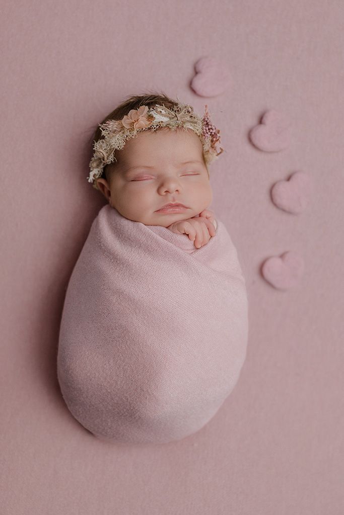 Newborn baby girl swaddled in pink wrap wearing floral headband, sleeping peacefully on pink background with heart decorations.