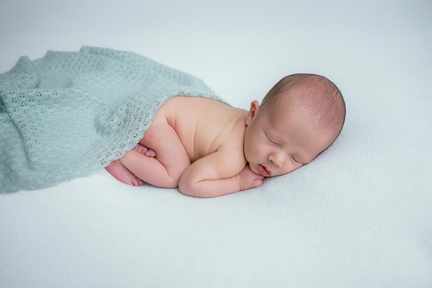 Newborn baby sleeping peacefully on white blanket, wrapped in soft teal knit wrap, curled in fetal position.