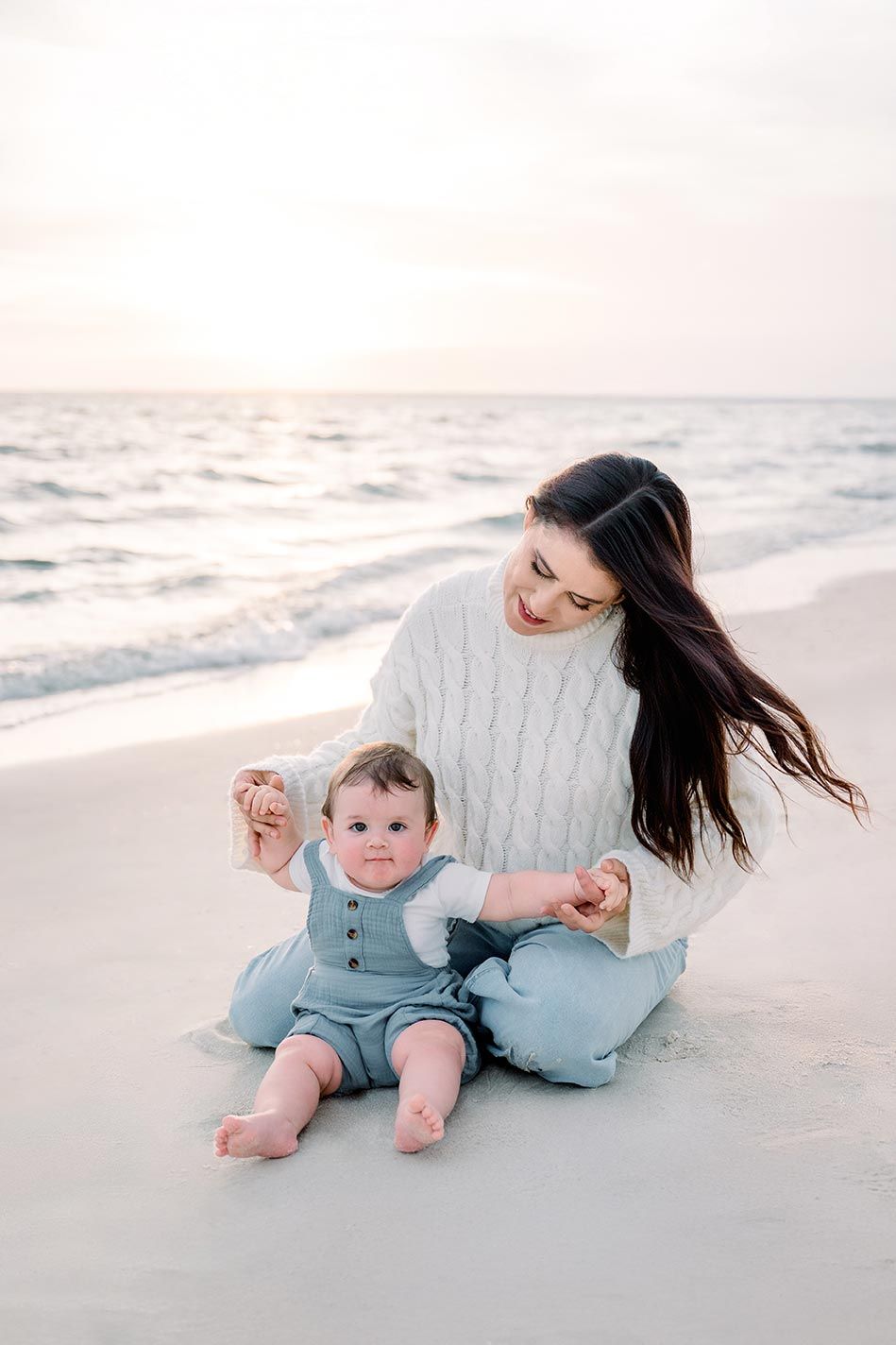 A mother sits on a sandy beach with her baby in a blue romper at sunset.