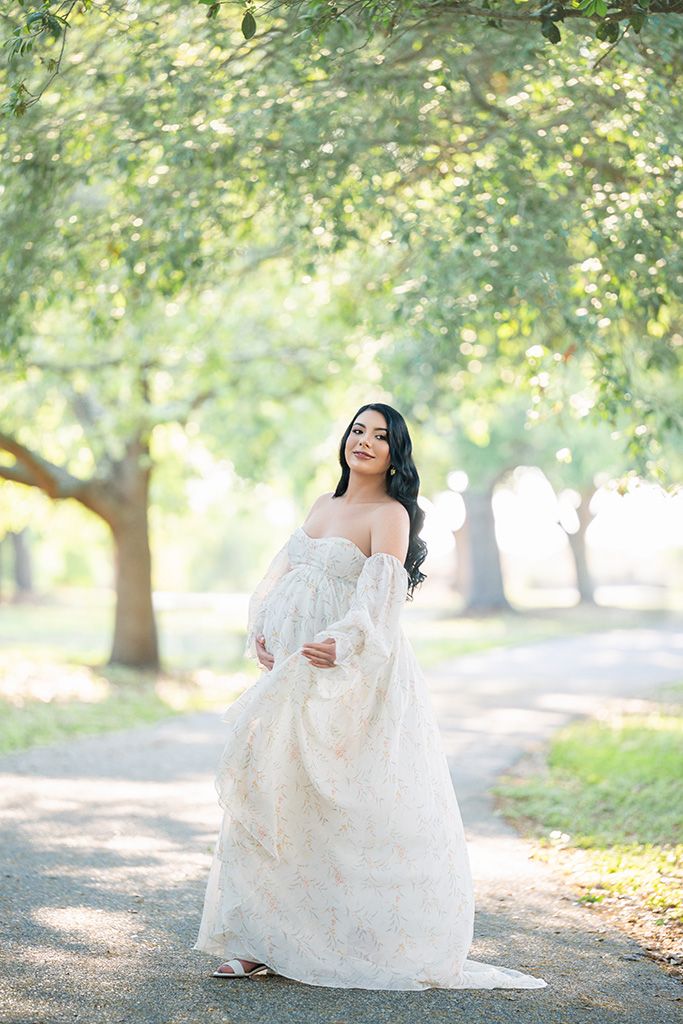 Pregnant woman in elegant off-shoulder white lace gown poses gracefully on a tree-lined path in a lush green park.
