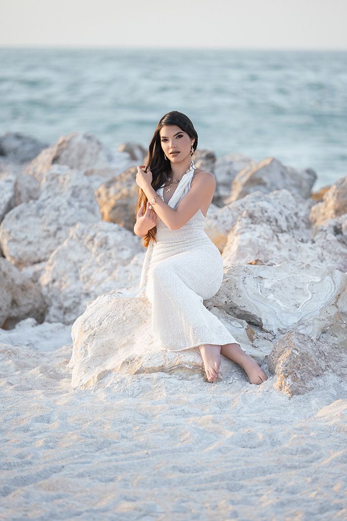 Elegant woman in white dress posing on rocky shoreline with ocean waves behind her.