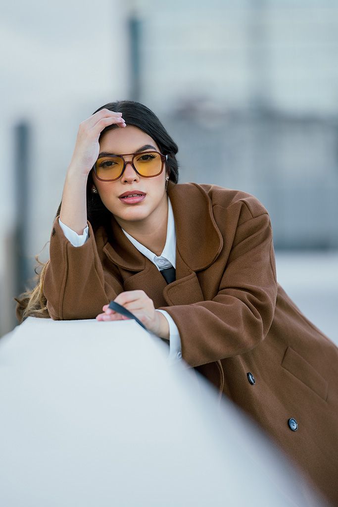 Close-up of brunette woman in camel coat and yellow sunglasses leaning on white ledge in urban setting.
