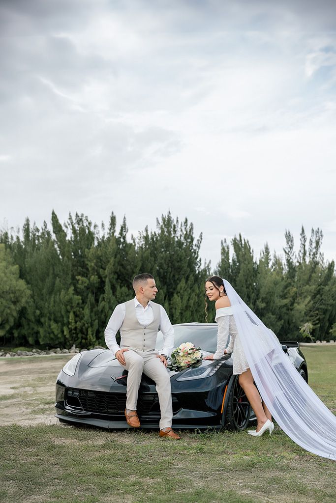 Bride and groom pose by a black sports car outdoors, surrounded by lush trees under a cloudy sky.