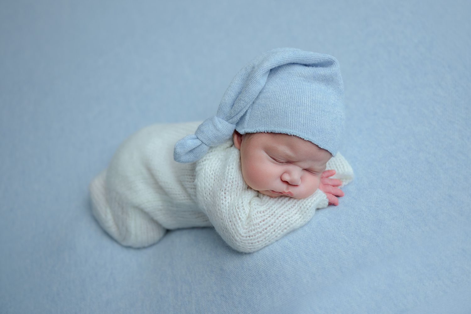 Newborn baby sleeping peacefully wrapped in white swaddle wearing a blue knot hat on a soft blue background.