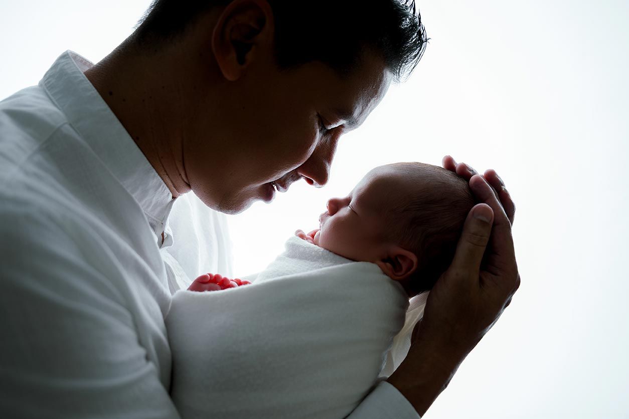 Father tenderly holds newborn baby in white blanket, sharing an intimate moment in soft backlit portrait.