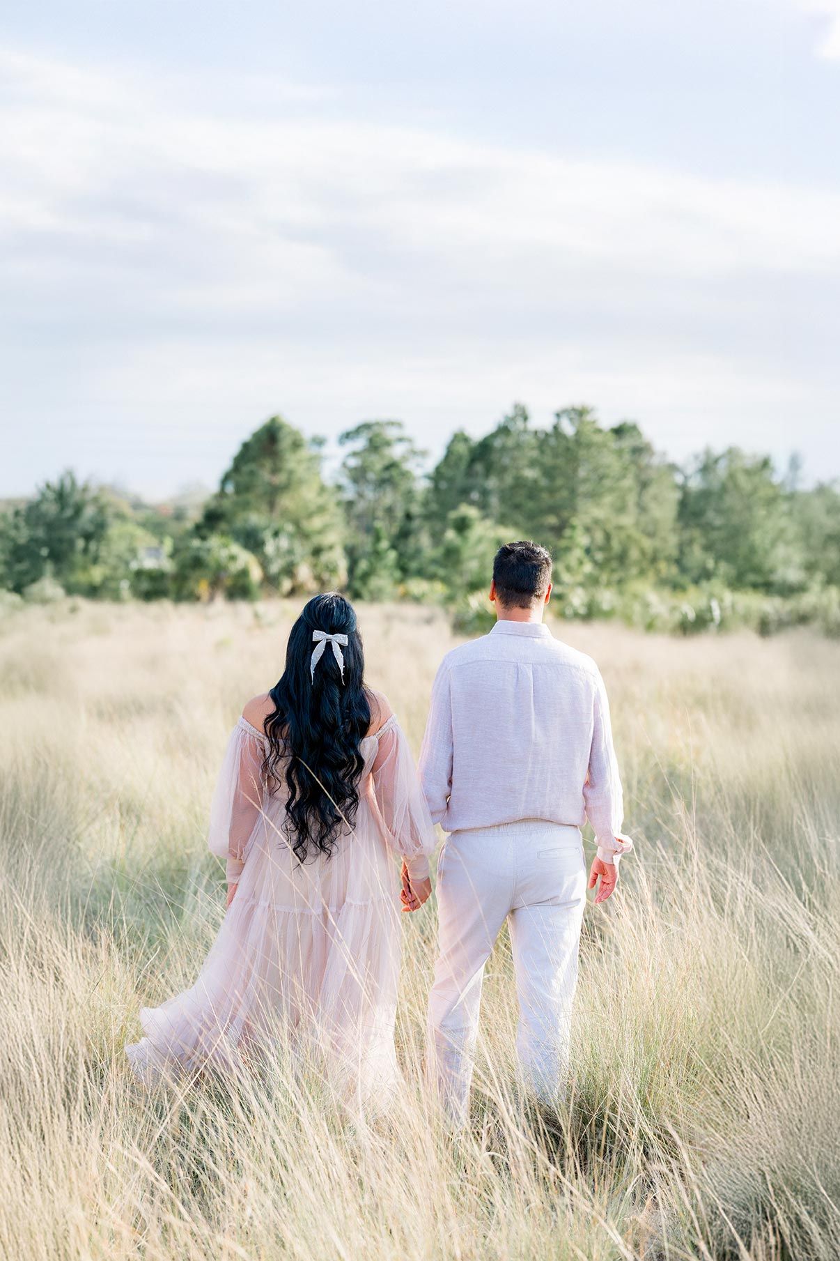 A couple walks hand in hand through tall grass fields, woman in pink gown with bow, man in white suit.