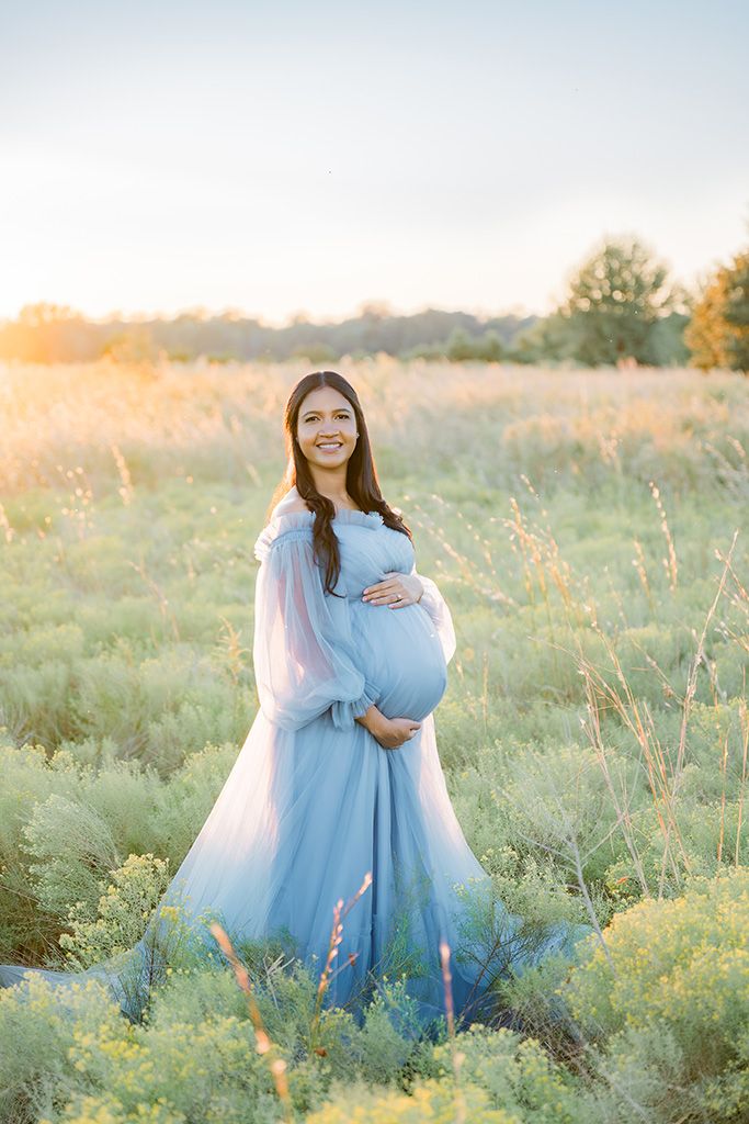 Smiling pregnant woman in a flowing blue gown cradling her belly in a sunlit field at golden hour.