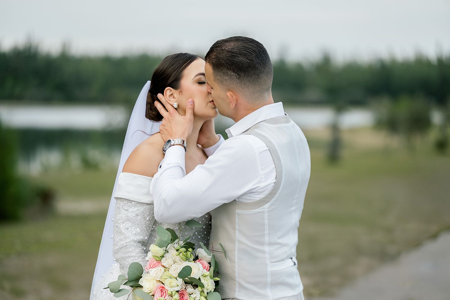 Bride and groom share a romantic kiss outdoors, bride holding a pink and white bouquet, with a scenic lake and trees behind.