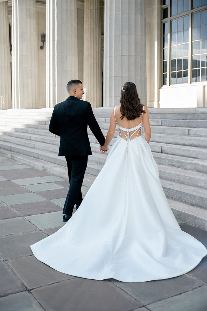Bride in white gown with open back and groom in black suit holding hands walking up stone steps.