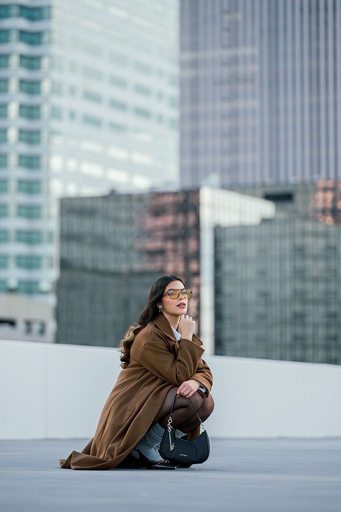 Elegant woman in brown coat and yellow sunglasses poses seated on urban rooftop with city skyscrapers behind her.