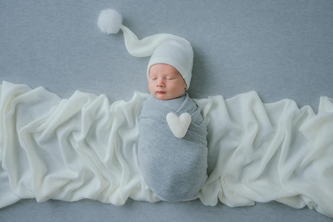 A sleeping newborn wrapped in a gray swaddle with a white heart, wearing a long white knit hat on a white fabric backdrop.