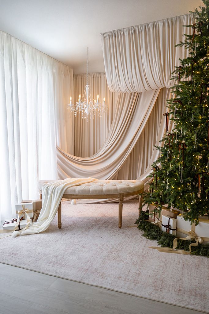 Elegant Christmas room with draped cream curtains, crystal chandelier, tufted bench, and decorated Christmas tree with gold accents.