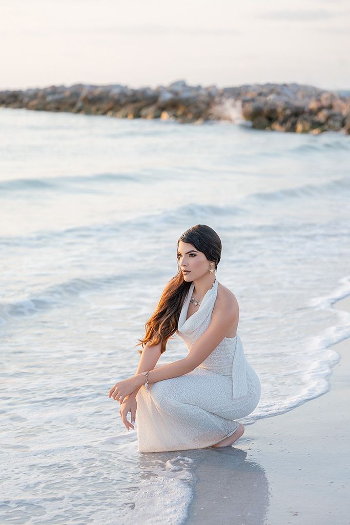 Brunette woman in white halter dress crouching gracefully at water's edge on beach.