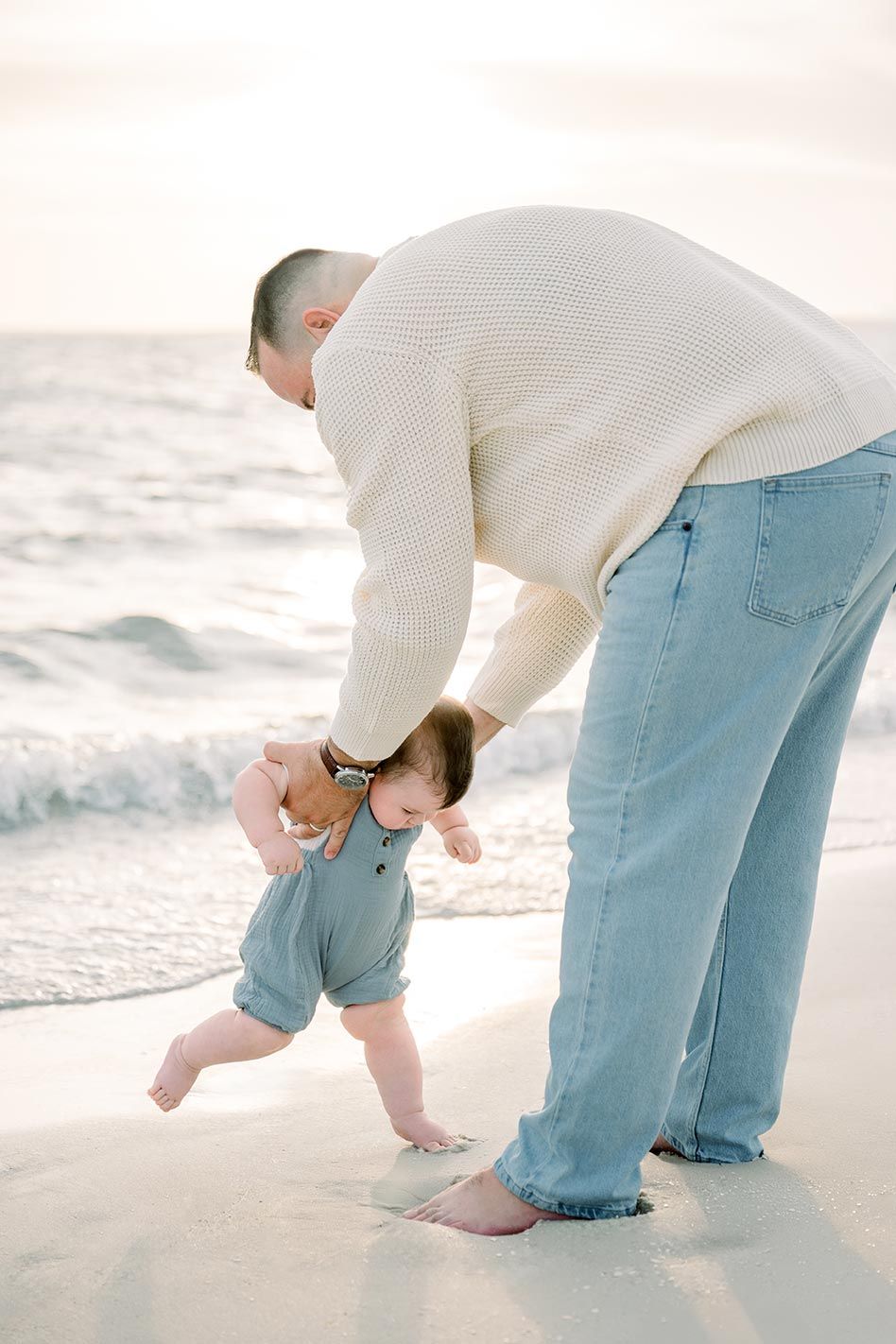 An adult holds a small child's hands as they walk together on a sandy beach at sunset.