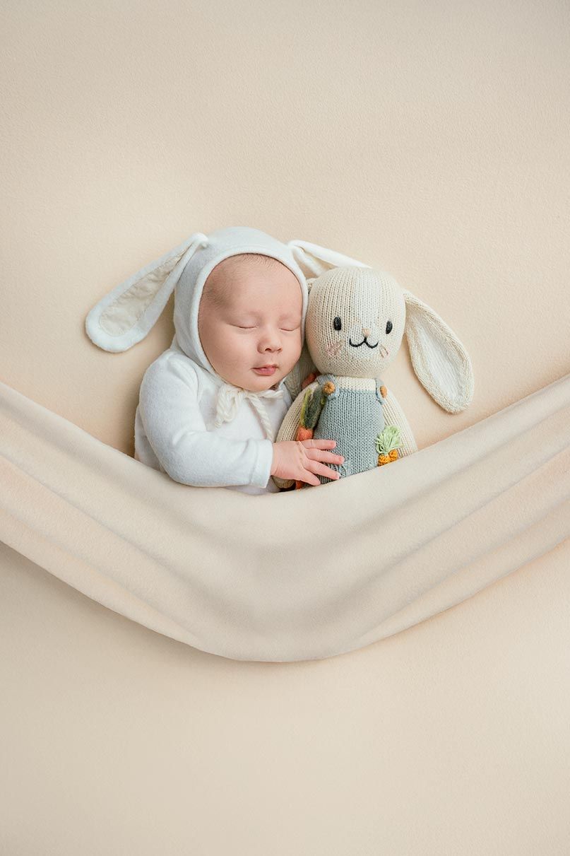 A baby in a white sweater sleeps peacefully in a cream-colored hammock with a plush bunny toy.