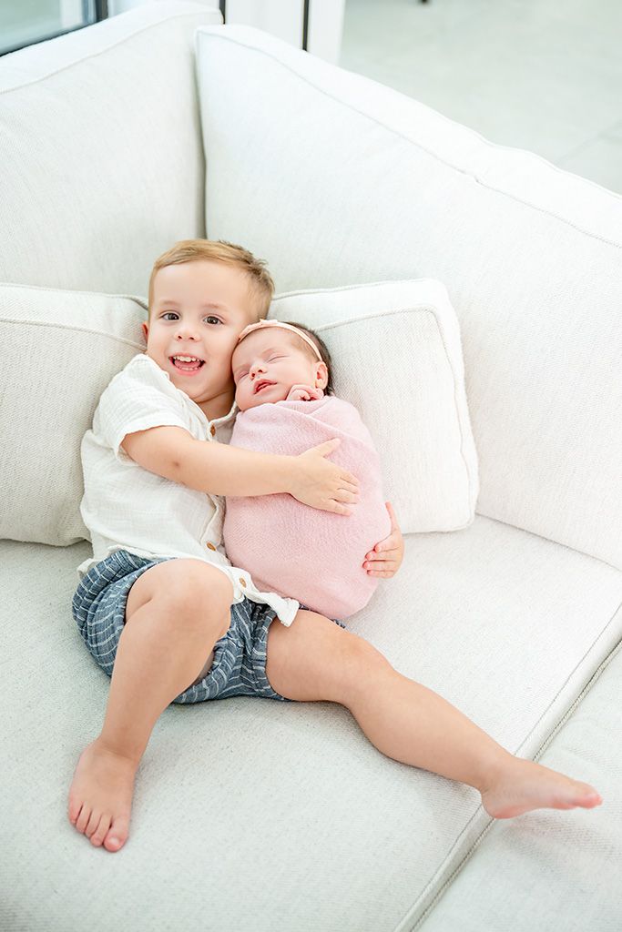 Young boy smiling on white sofa holding newborn baby sibling wrapped in pink blanket.