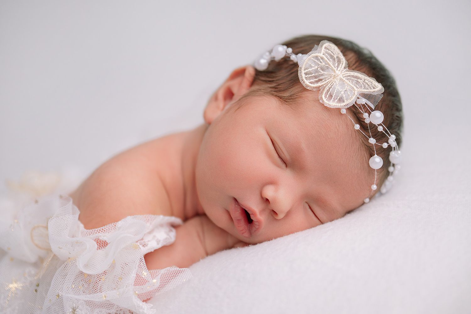 Peaceful newborn baby sleeping with pearl butterfly headband on white fluffy blanket in professional photo.