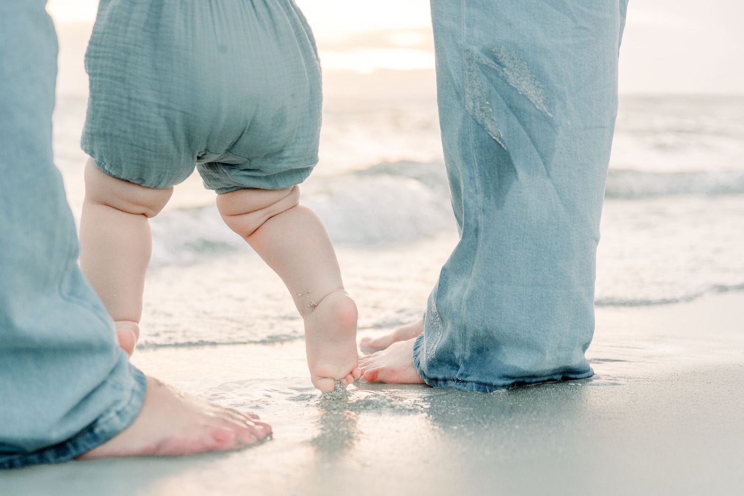 Bare feet and legs of parent and baby standing together on a sandy beach by the ocean waves.