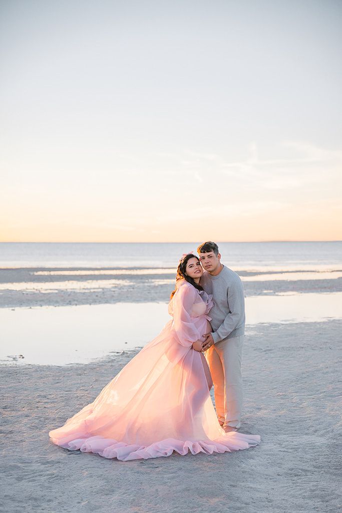 Couple embracing on a beach at sunset, woman in flowing pink gown, man in gray, with calm ocean waters behind them.