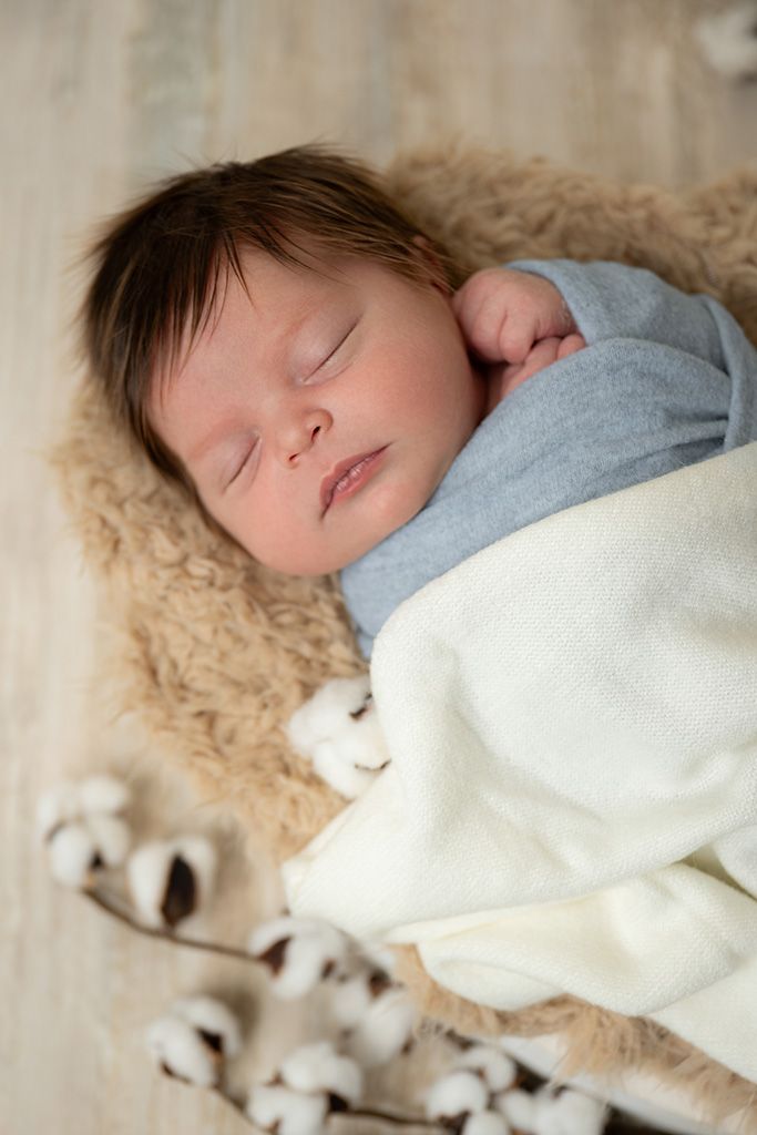 Peaceful newborn baby sleeping on a fluffy beige fur blanket, wrapped in a soft blue and white swaddle with cotton stems.