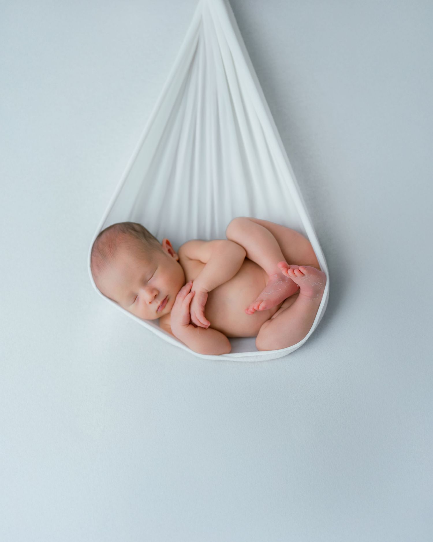 Peaceful newborn baby sleeping curled up in a white fabric hammock against a soft blue background.