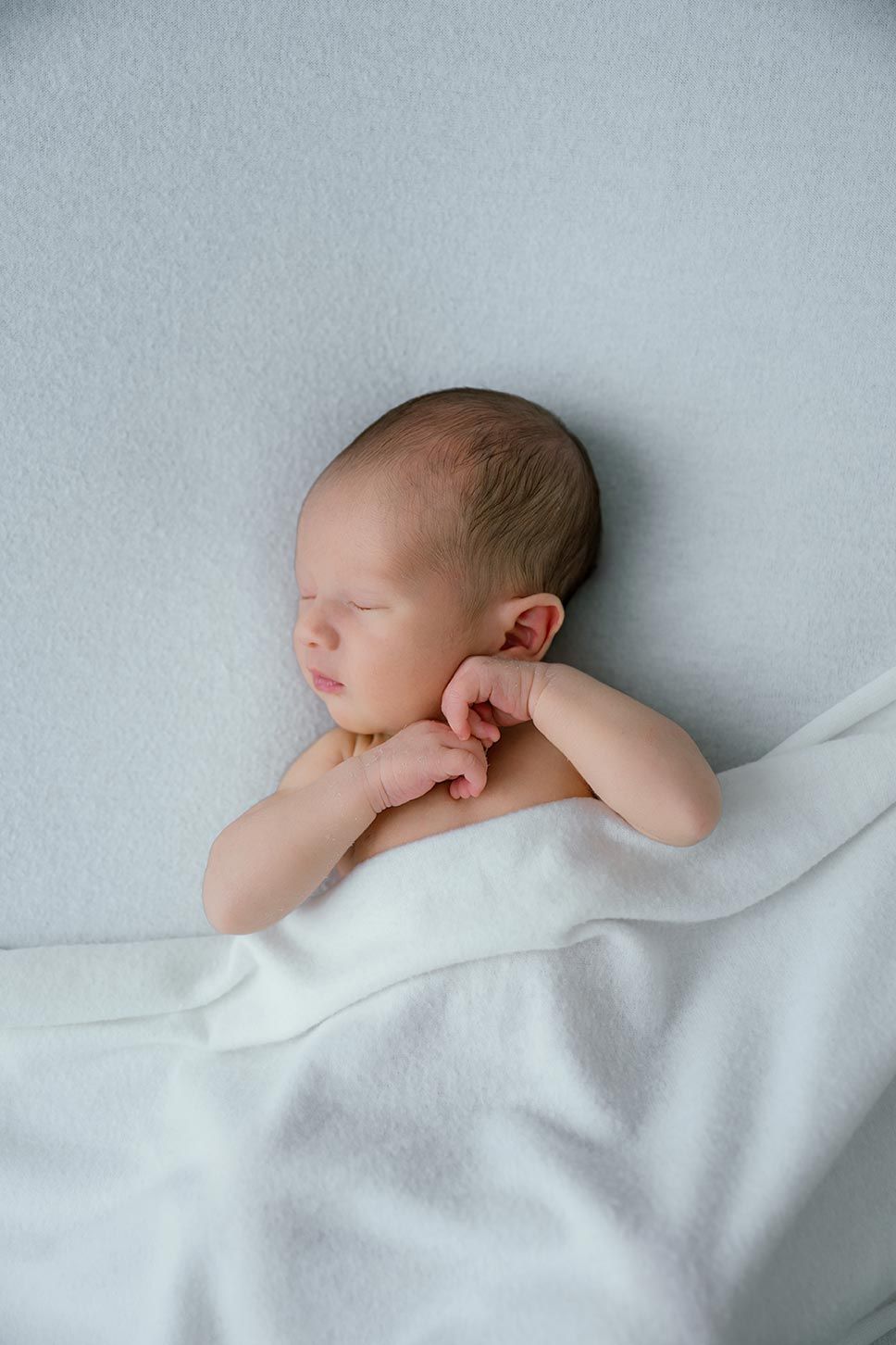 A sleeping newborn baby wrapped in soft white blankets rests peacefully against a light grey background.