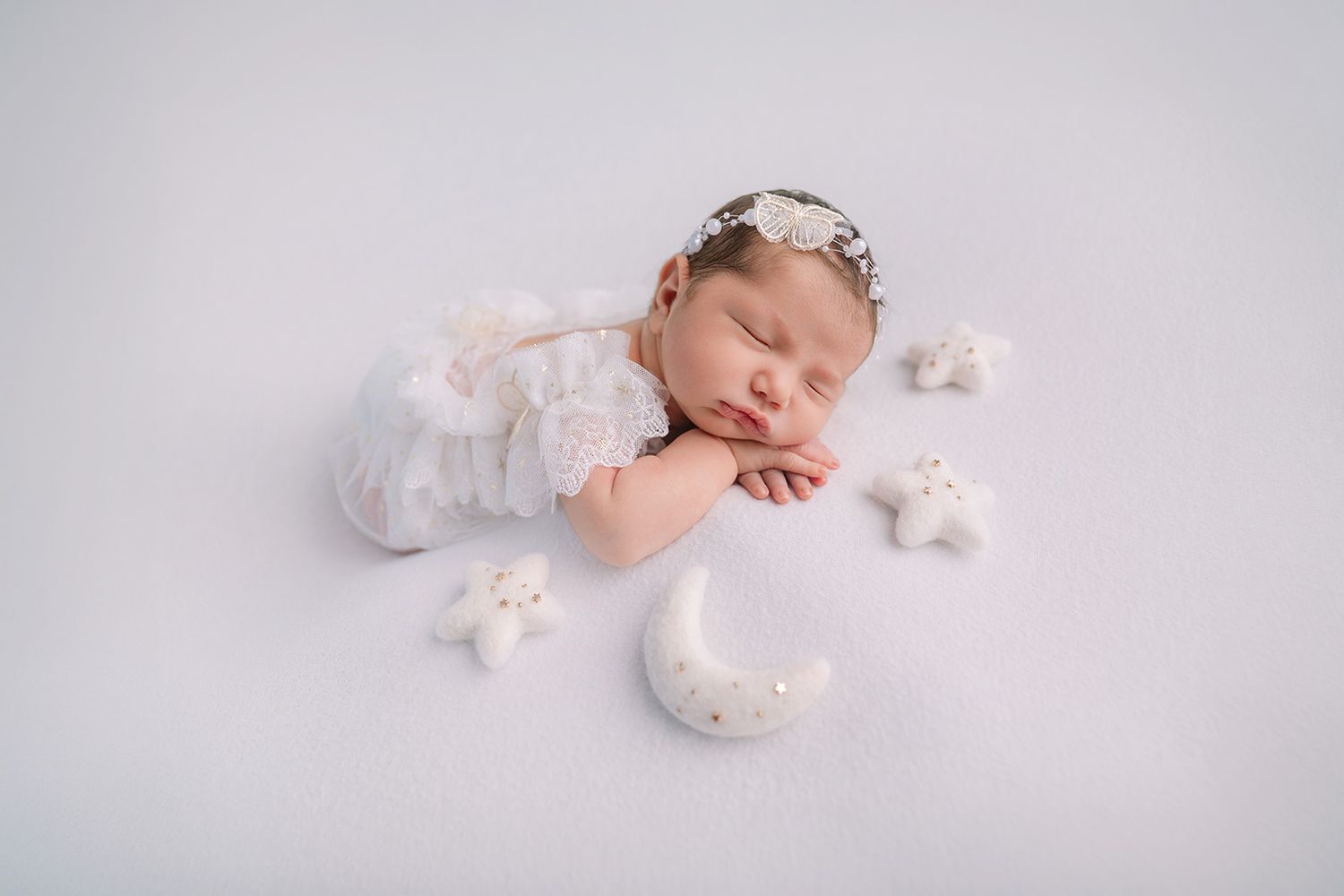 Sleeping newborn baby in white lace outfit with floral headband surrounded by star and moon props on white background.