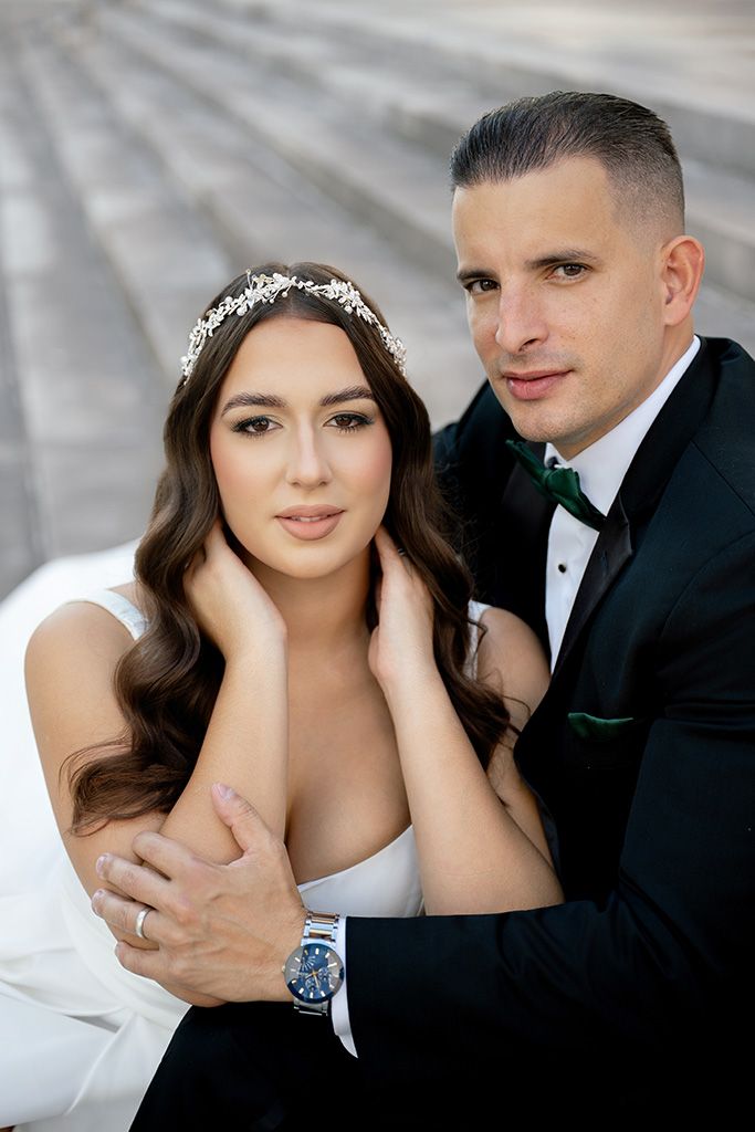 Bride and groom sitting on steps, groom embracing bride wearing white gown and floral crown.