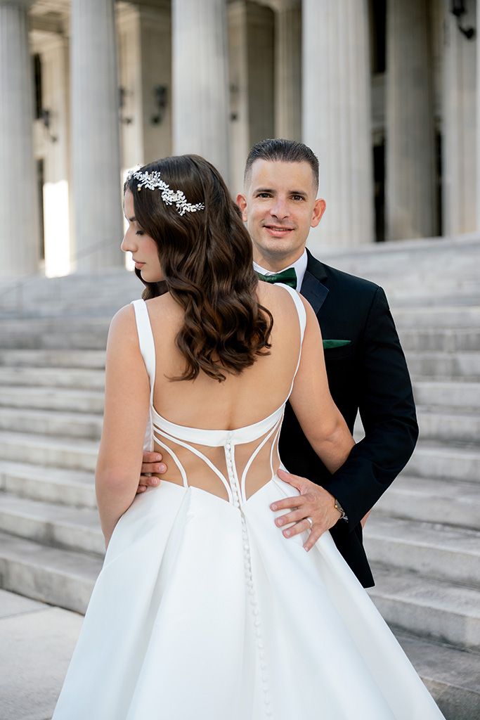 Bride in backless white gown with groom in black tuxedo posing on grand steps with columns.