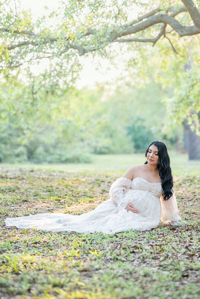 Pregnant woman in white lace gown sitting gracefully under trees in a sunlit park during maternity photo session.