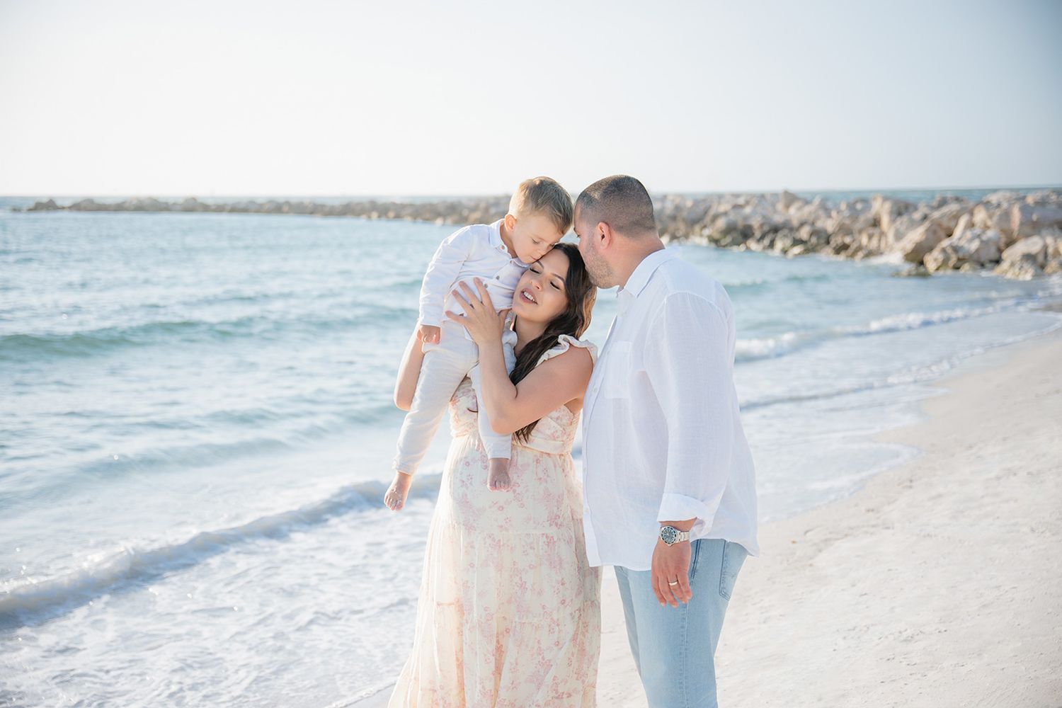 Loving family sharing a joyful moment on a scenic beach, parents kissing child between them near rocky shore.