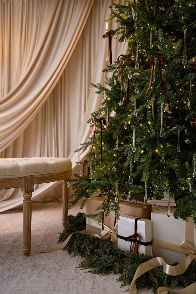 Elegant Christmas tree with crystal ornaments and warm lights, gifts underneath, beside a cream bench and draped curtains.