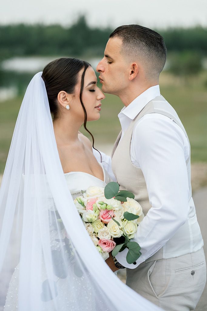 Groom kisses bride on forehead outdoors, bride holding pink and white rose bouquet in wedding attire.