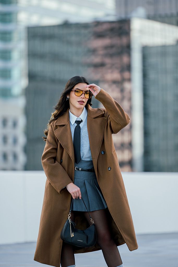 Woman in camel coat, gray skirt, tie, and amber sunglasses holding a navy bag in an urban setting.