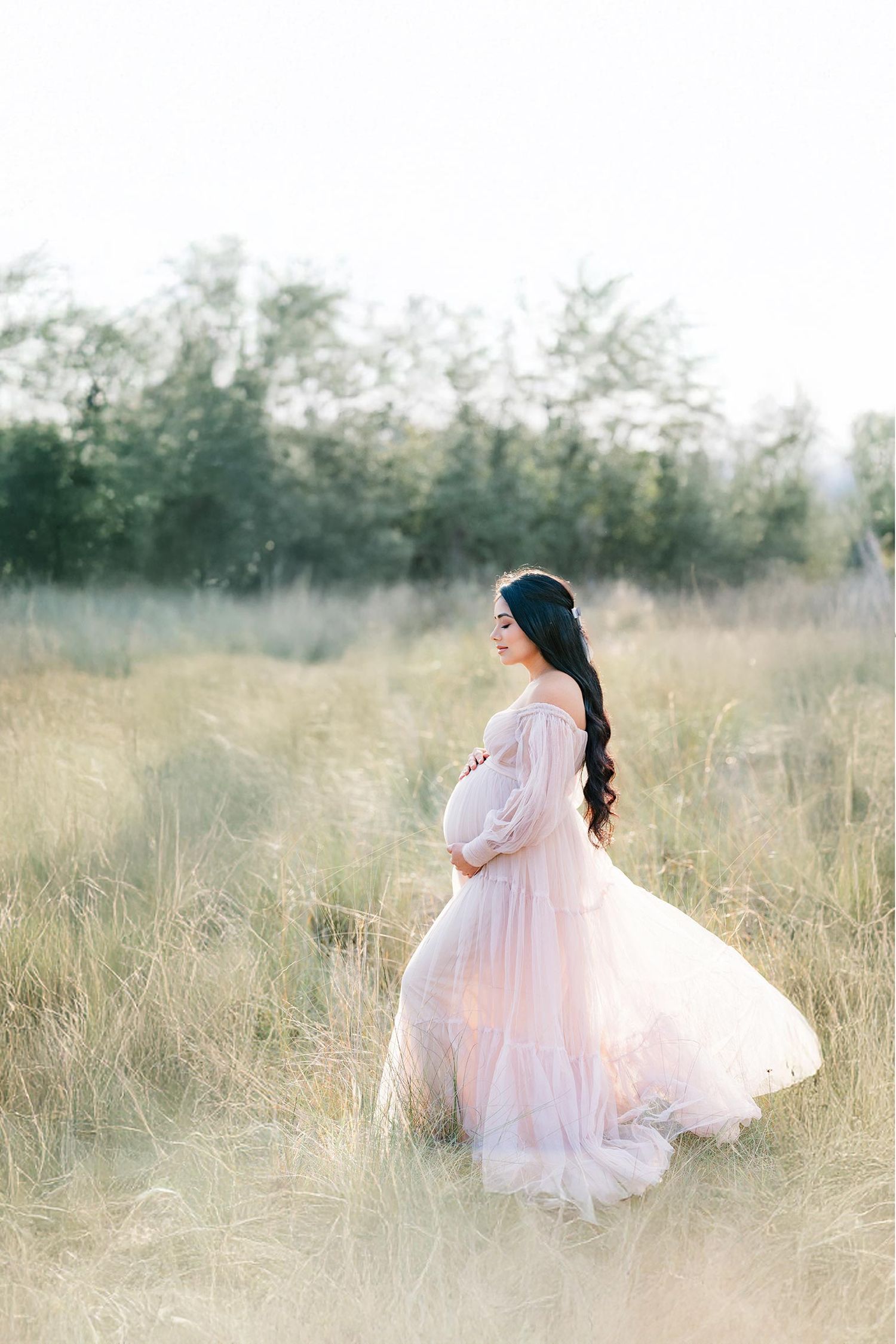 Expectant mother in flowing pink dress twirls gracefully in sunlit meadow with tall grass and trees in background.