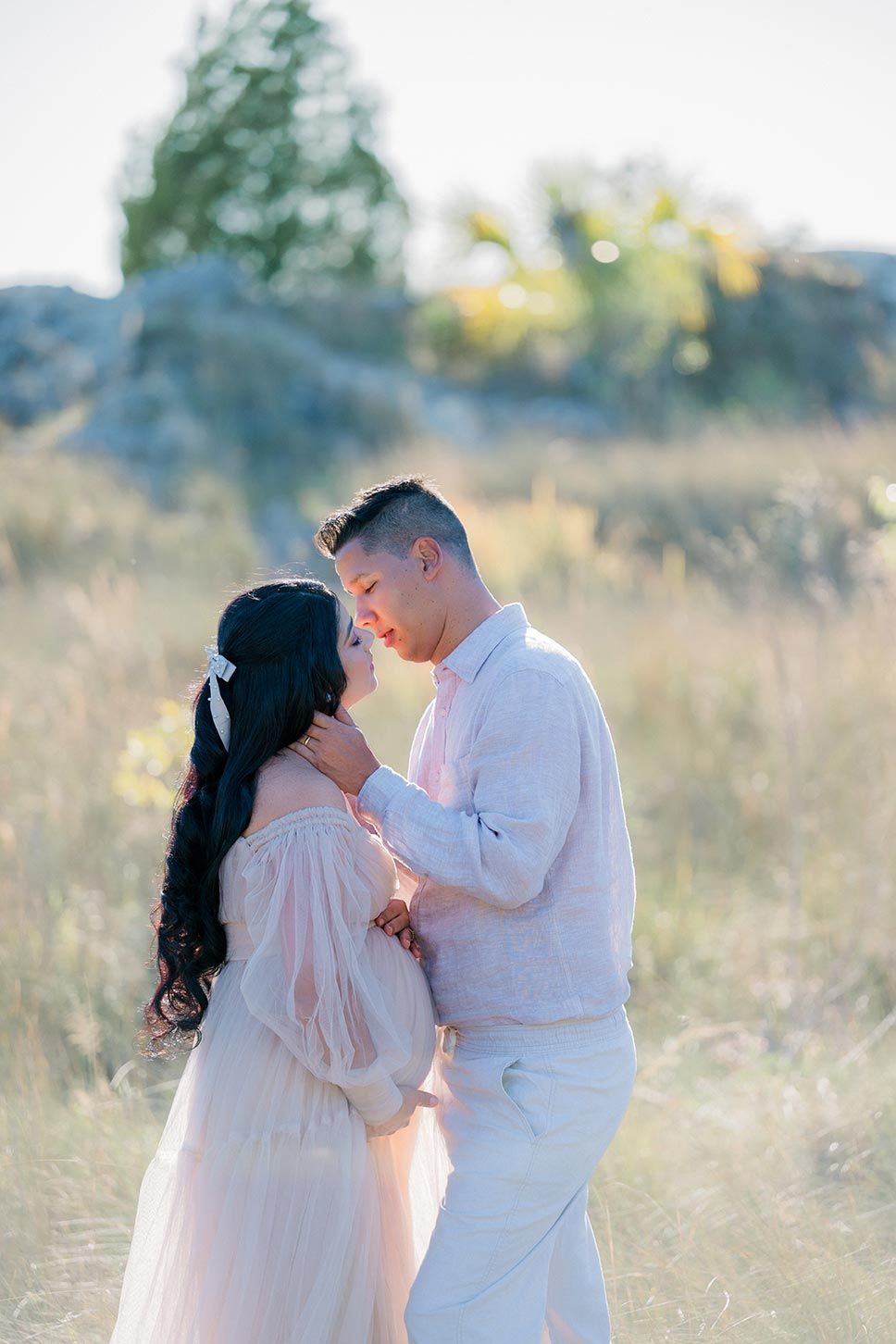 A romantic couple in white attire embracing in a sun-lit meadow during a dreamy outdoor photo session.