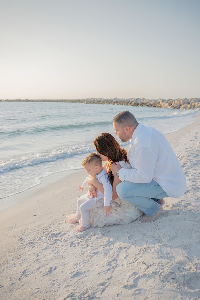 Family with toddler crouching together on white sand beach at sunset.