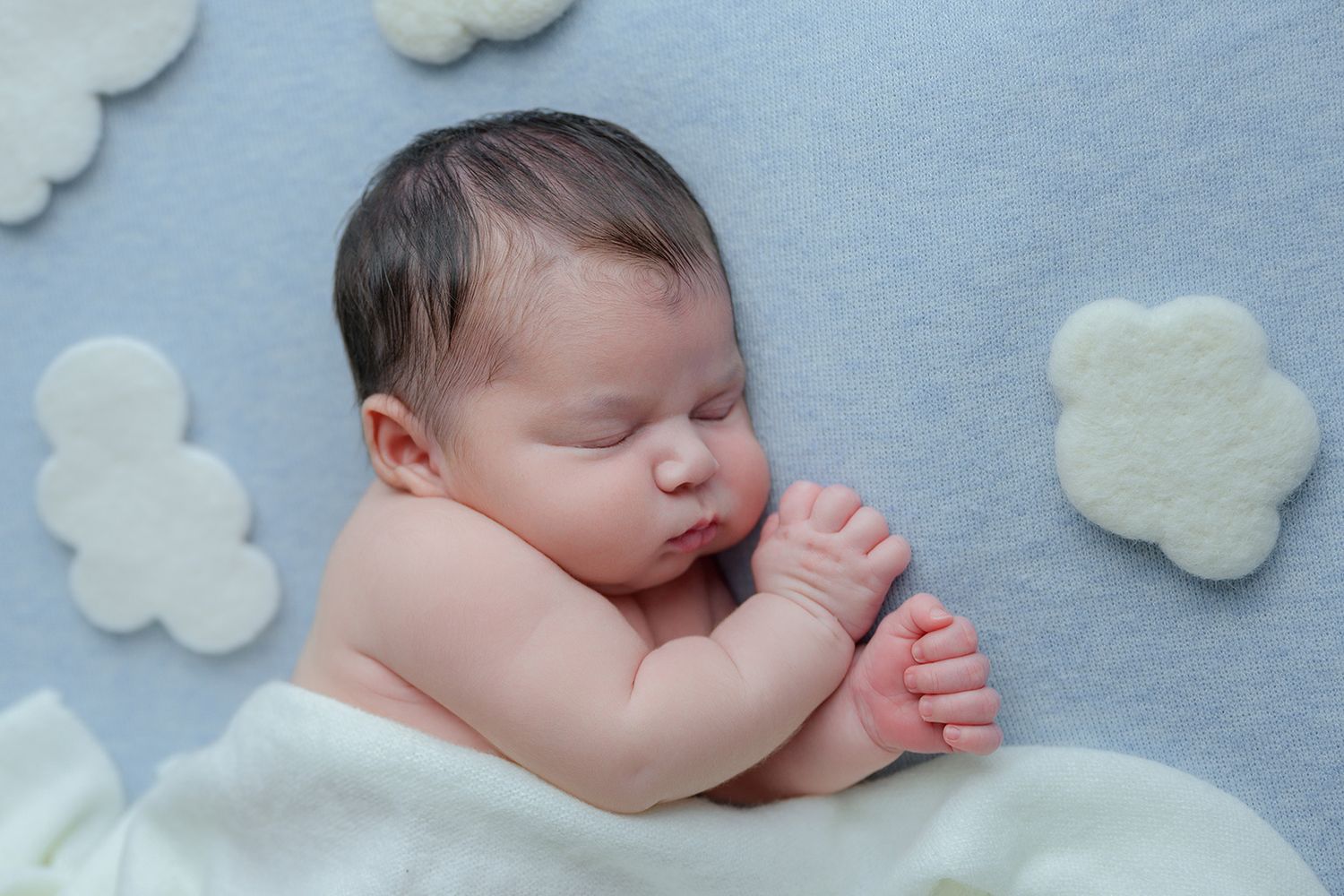Peaceful newborn baby sleeping on a soft blue blanket surrounded by white felt cloud decorations.