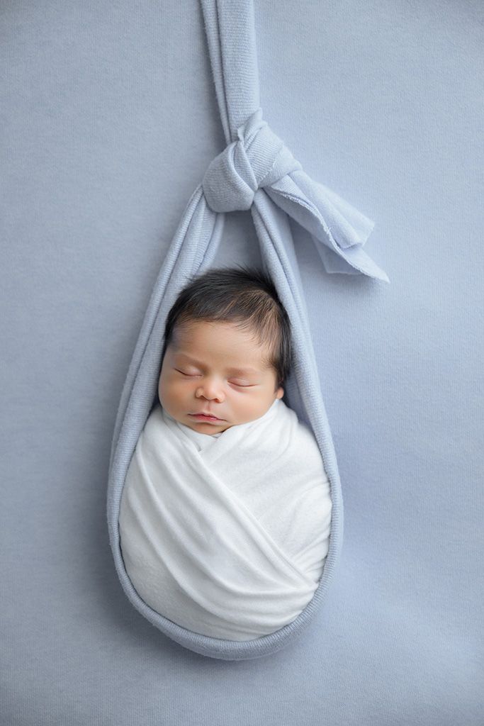 Newborn baby swaddled in white wrap resting in a blue fabric sling prop.