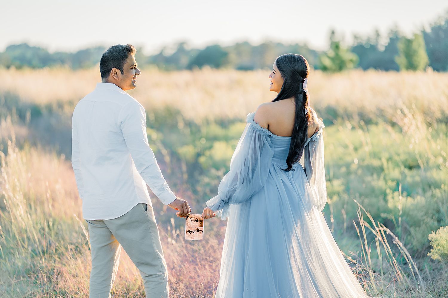 Couple holding hands in a golden field, woman in blue gown, man in white shirt, romantic outdoor photo shoot.