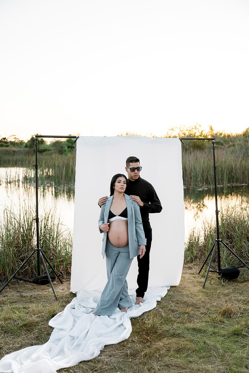 Outdoor maternity photo shoot near a pond with white backdrop and couple silhouetted against natural scenery at sunset.