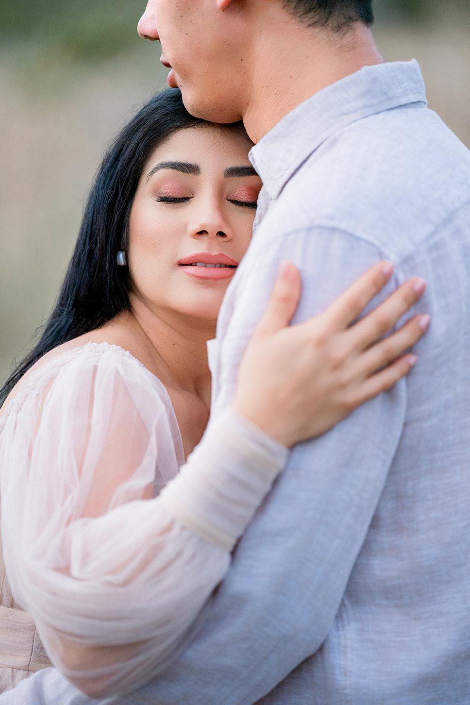 A romantic couple embraces tenderly outdoors, with the woman in a soft pink blouse resting against her partner in grey.