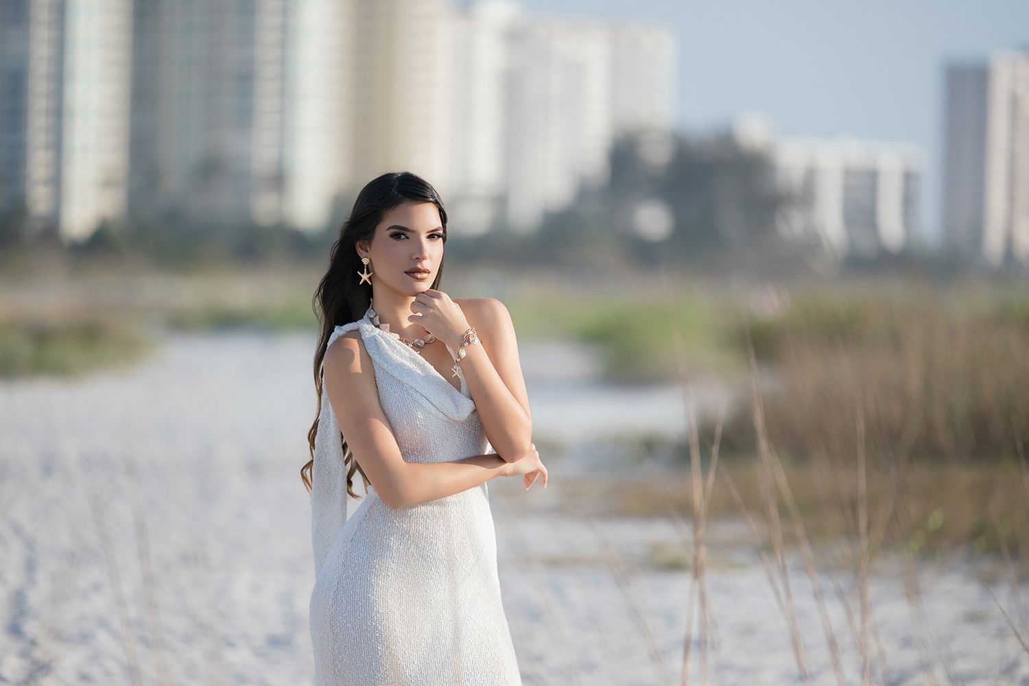 Elegant brunette woman in a white lace dress posing on a sandy beach with city buildings in the background.